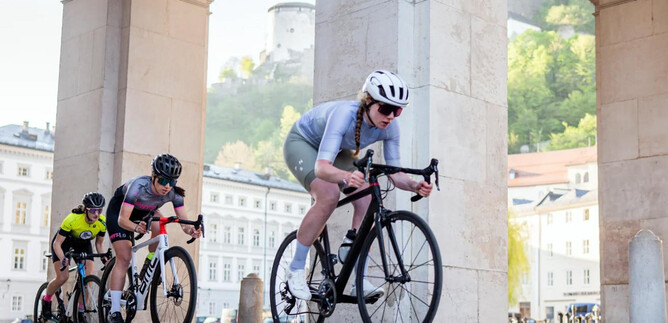 Drei Radfahrer fahren unter einem Bogen hindurch. Die Landschaft im Hintergrund zeigt eine Stadt mit grünen Hügeln und historischen Gebäuden. | © Cyclodom